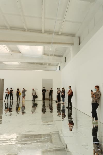 A large white room with a mirrored floor. People stand around the outside, taking photographs and exploring the space.
