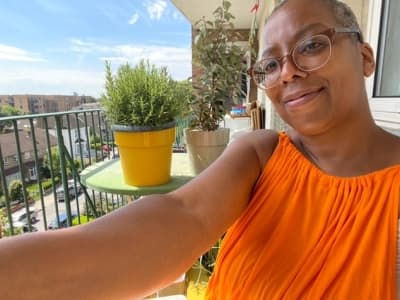 Gaylene is to the right of the image sitting on her balcony smiling, she is wearing an orange sleeveless top. To the left of an image are two plants, one in a yellow pot and one in a cream pot on top of a green table and balcony rails. There is blue sky in the background with some buildings.
