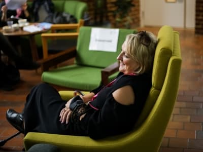 Image description: A woman with shoulder length blonde hair sits sideways in a lime green arm chair, her hands are crossed on her lap, she isn't looking at the camera.