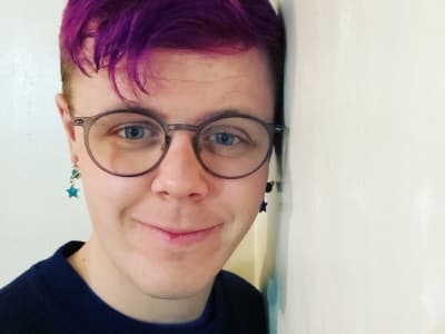 A white person with brown round glasses, star earrings, blue eyes and purple hair.