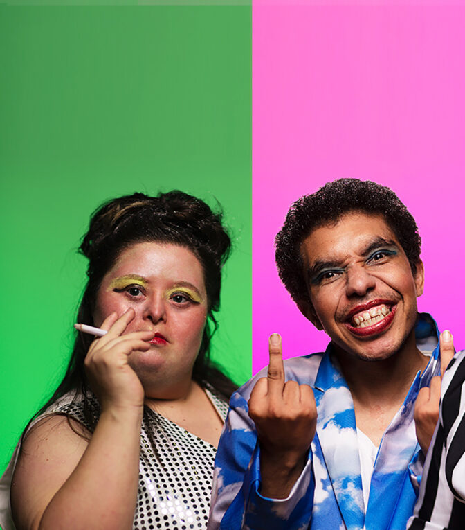 Two heavily made-up individuals with a pink and green background. One is raising their middle finger.