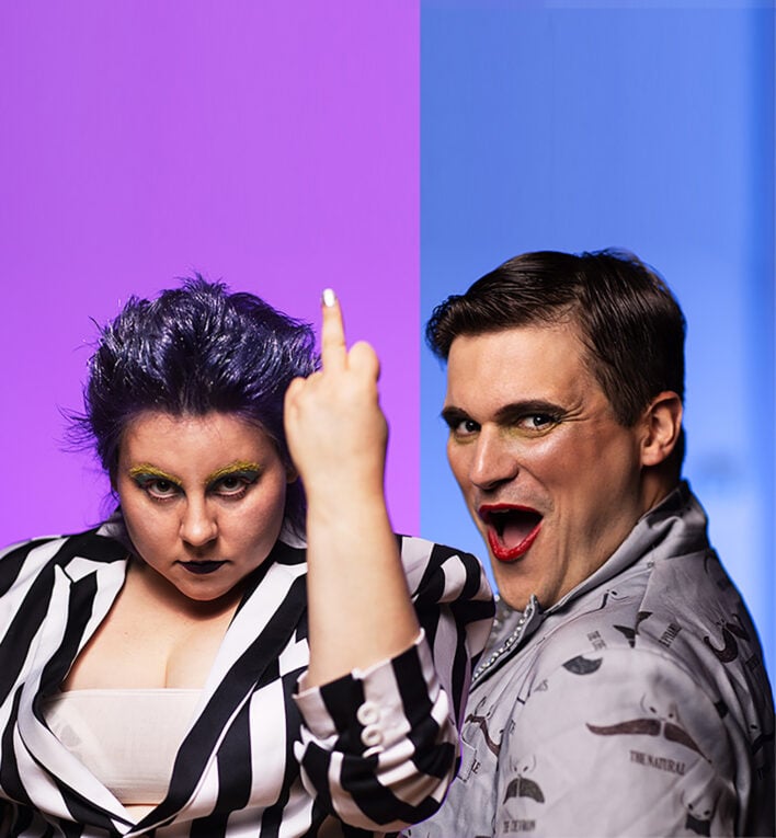 Two heavily made-up individuals with a purple and blue background. One is raising their middle finger.