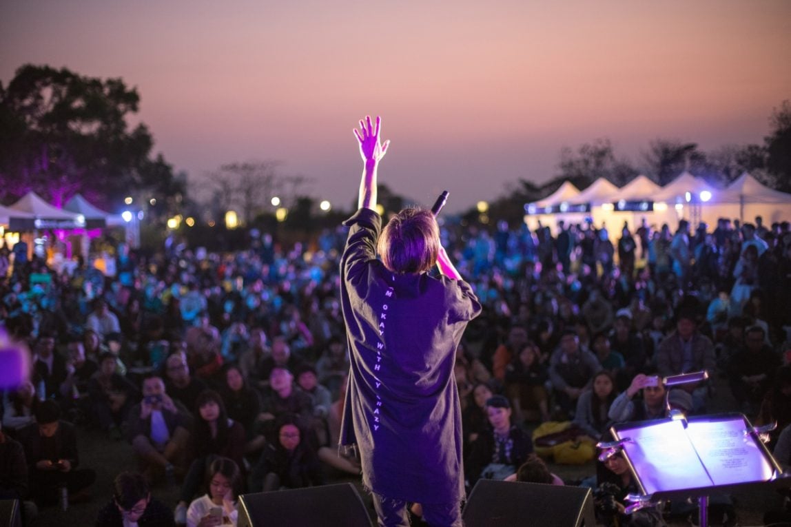 A performer on stage singing with their arm raised, it is night time and there is a large audience