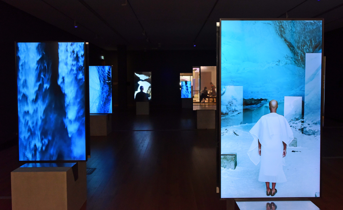 A gallery installation in a dark room, there are a series of tall screens showing animated scenes