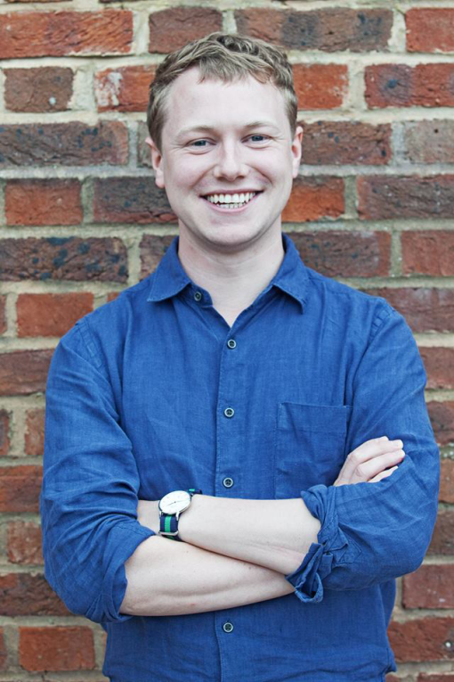 A headshot of a person standing in front of a brick wall wearing a blue shirt
