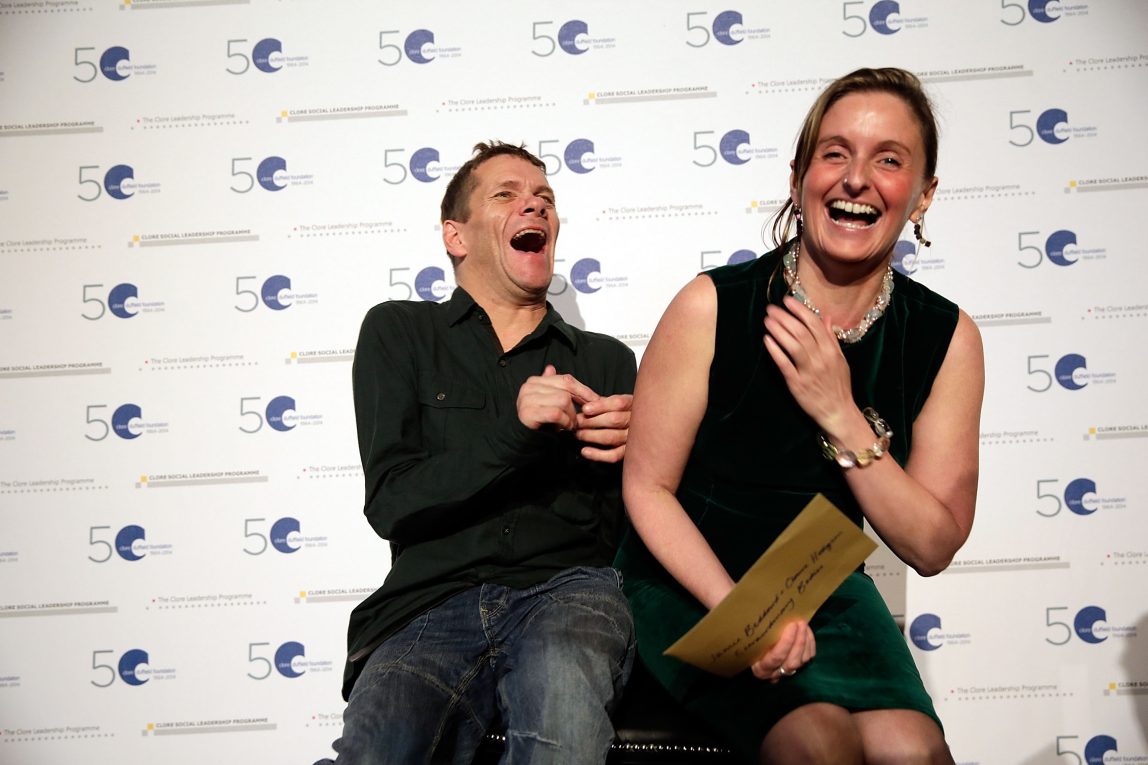 Two people sat on a single chair and laughing in front of a step and repeat