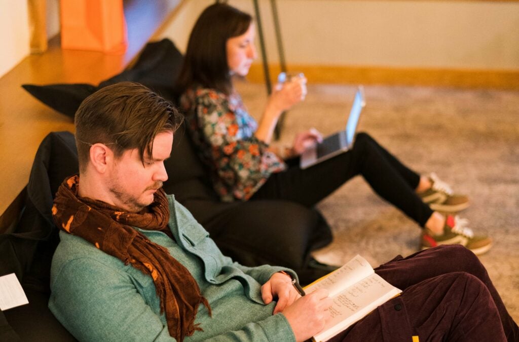 Two fellows sitting on the floor on bean bags, one writes in a notebook, the other types on a laptop