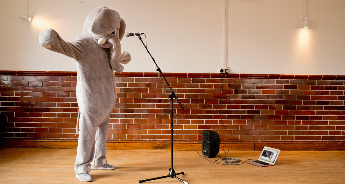 Person dressed in an ill fitting elephant suit standing in front of a microphone. 