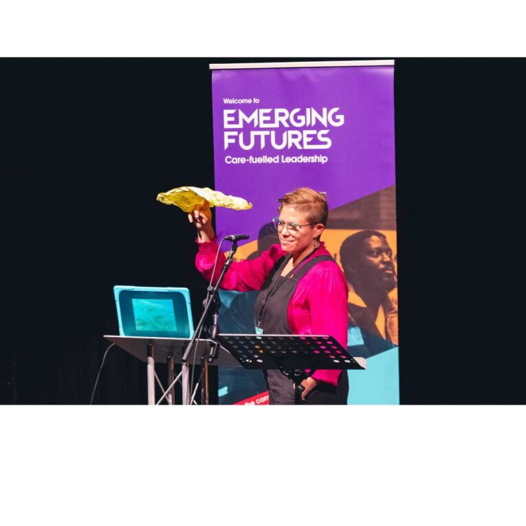 A speaker on stage at the Emerging Futures 2023 conference holding a piece of paper which had been scrunched up and flattened back out