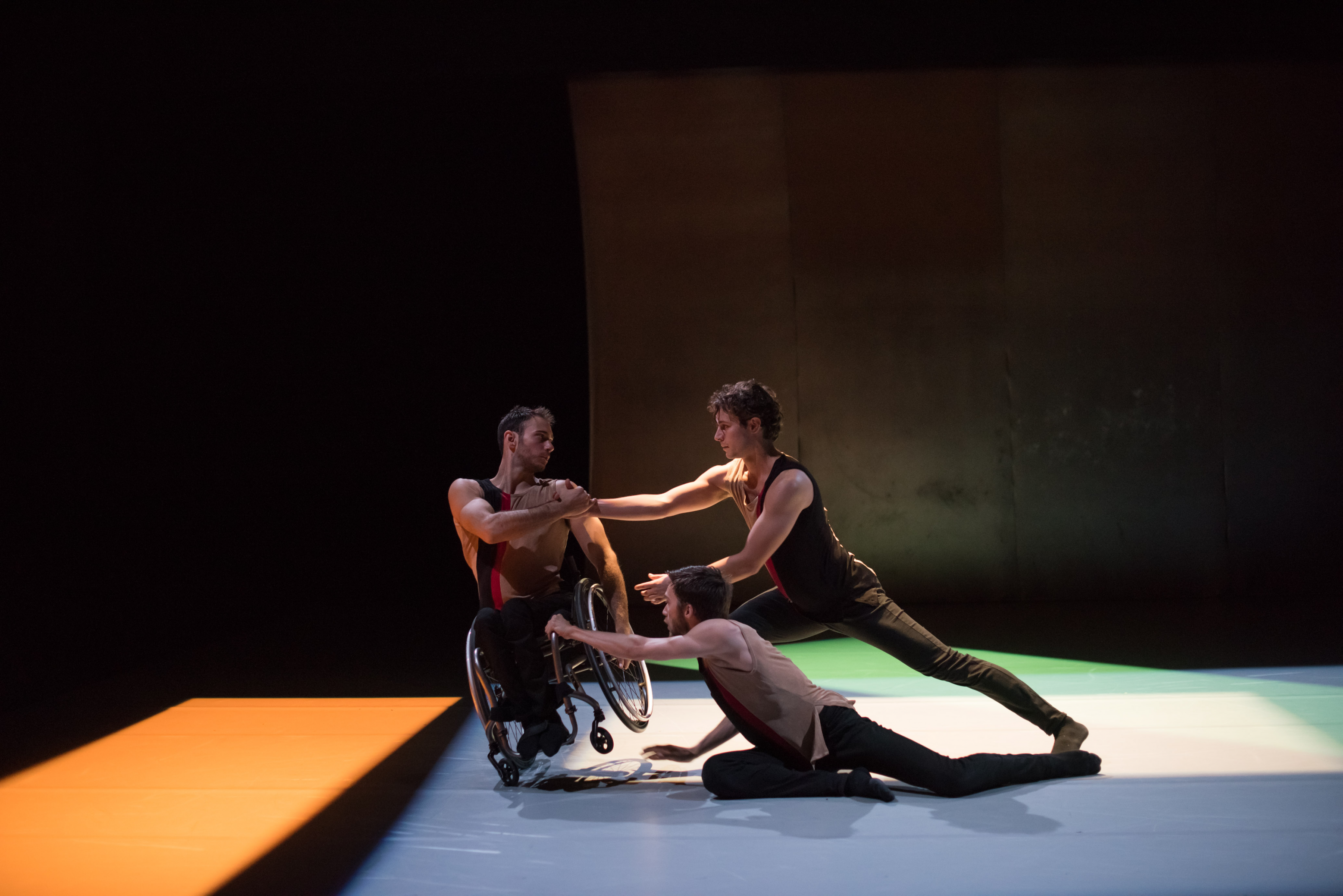 A troupe of three dancers support each other's bodies in a move on stage; one of the dancers is a wheelchair user. 