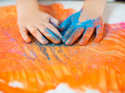 Two hands painting with orange paint and blue sand