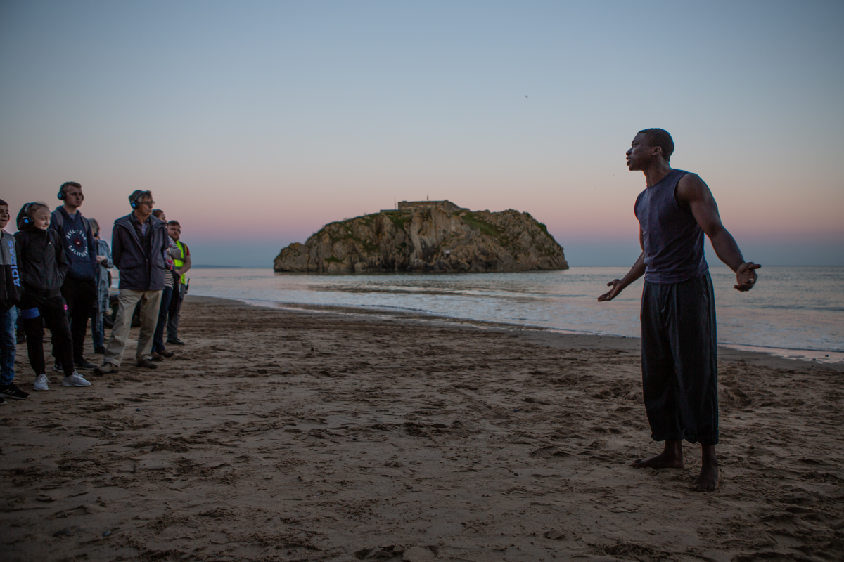 National Theatre Wales. Tide Whisperer. Photo by Jennie Caldwell a group of people with headphone on stood on a beach in front of a person who is speaking