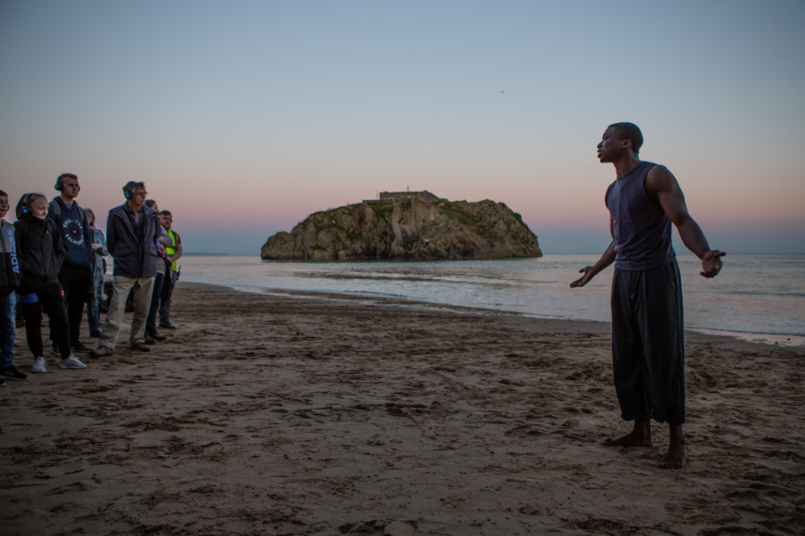 a group of people with headphone on stood on a beach in front of a person who is speaking
