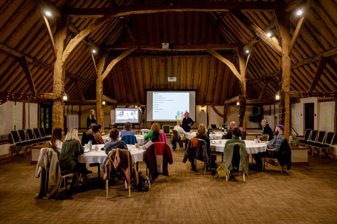 A group of fellows participating in a session, some are sat around round tables, some attend virtually via zoom