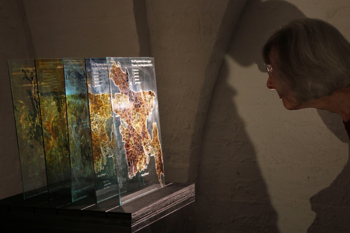 Stephen Bennett’s Layers of Bangladesh (2021), viewed by a participant at Glass House. A woman with cropped grey hair examining an exhibit made up of layers of maps on a plinth.