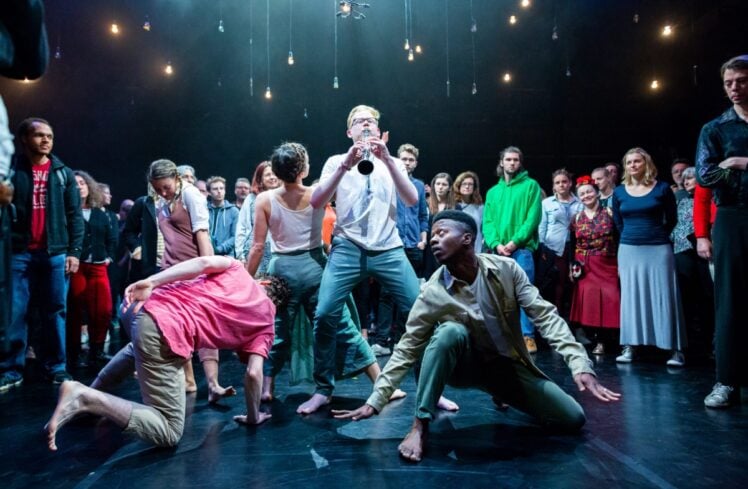 A group of performers stood in the middle of a crowd. One person in the middle is playing a clarinet and the rest dance around them
