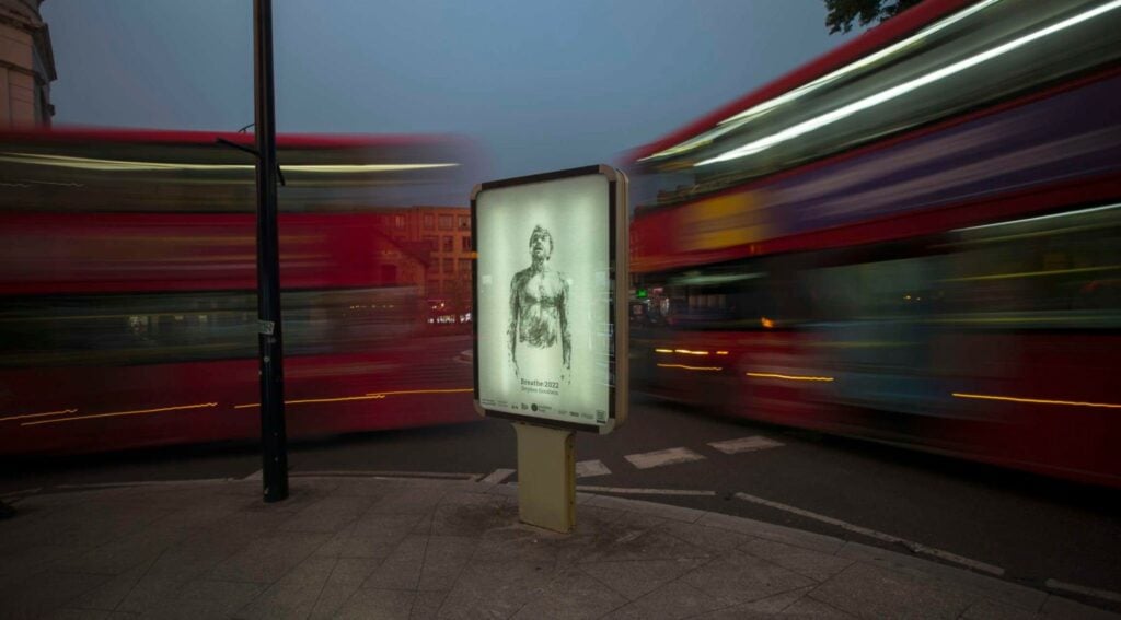 A long exposure photograph of an advertising sign showing a black and white illustration of a man, behind are two red London buses caught in motion. 