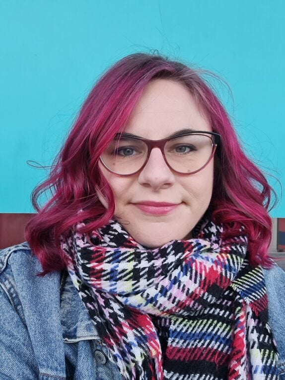 A mid 30’s person wearing glasses with mid length electric pink hair, contrasted expertly with a bright teal background – actually the DLR station.