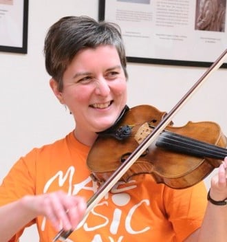 Picture of Alison Reeves, a white woman with short grey hair and an orange shirt, playing the violin and smiling.