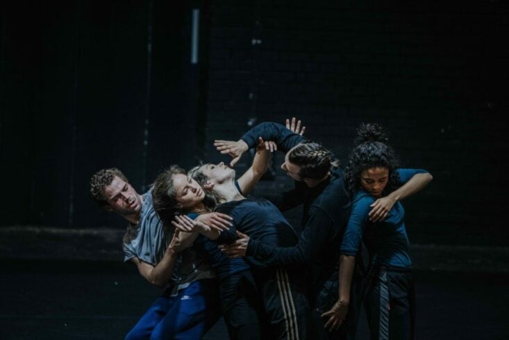 A group of dancers in the middle of a performance, they are gathered together against a dark background.