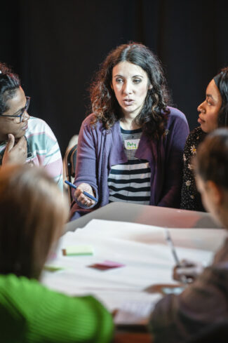 A table surrounded by a group of interested learners, headed by Paty Bennet - a woman with dark, curly hair and a purple cardigan.