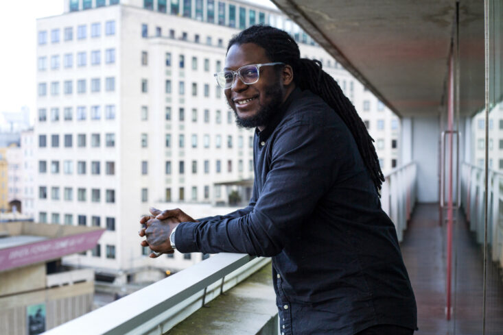 Portrait of Anthony Olanipekun smiling and leaning on a balcony of an apartment building.