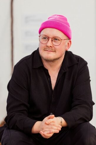 Relaxed photo of artist Garth Gatrix sitting wearing a pink hat, glasses and black shirt