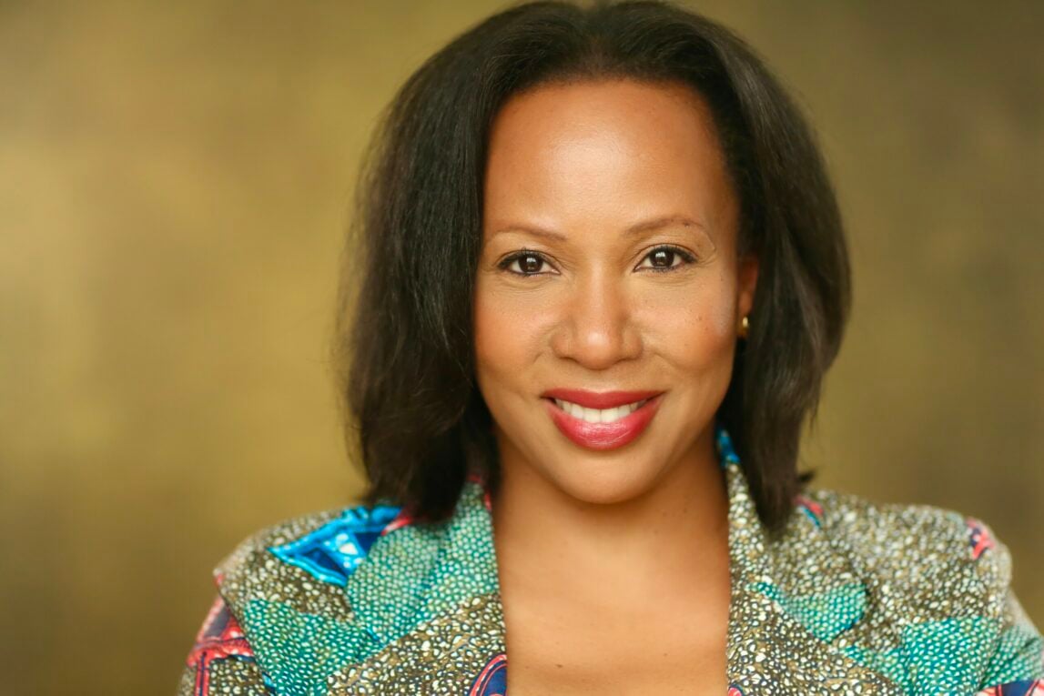 A Black woman with shoulder length hair, she is wearing red lipstick and smiling at the camera against a beige background.