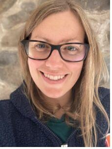 An informal profile picture of a white woman in her mid-30s with long dark blonde hair wearing a navy hooded fleece and big square tortoiseshell glasses smiling at camera. Behind her is a stone wall backdrop.