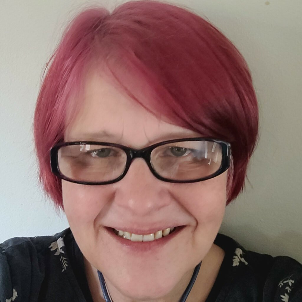Headshot of Liz Grren, she is wearing glasses and has short pink hair