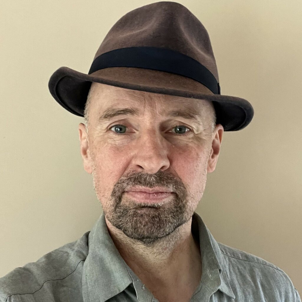 Headshot of David Jubb, he has a beard and is wearing a brown trilby hat 