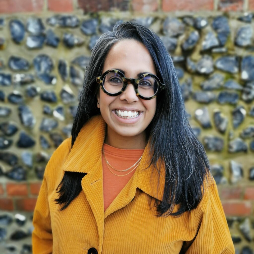 An Indian woman with shoulder-length salt-and-pepper hair, wearing round tortoiseshell glasses, gold chains, an orange top with a mustard jacket, is smiling warmly.