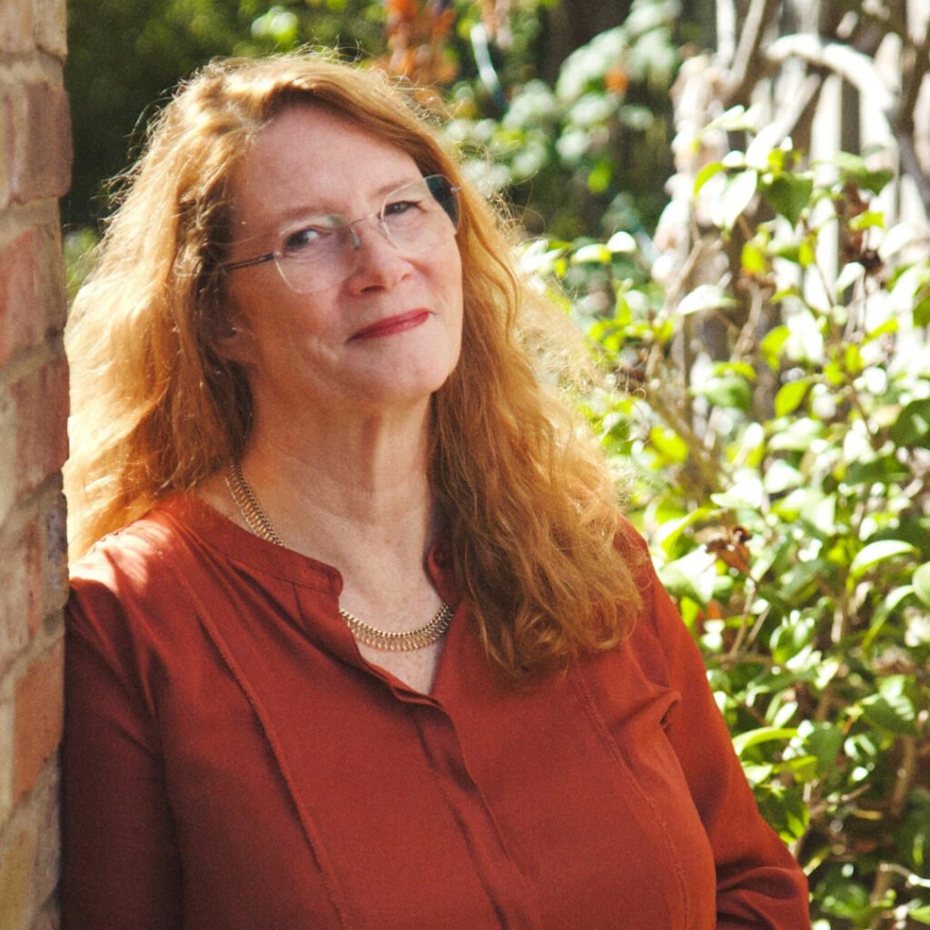 Headshot of Hedda Archbold, she is stood outside wearing a red shirt and glasses. She has long red hair and is smiling.