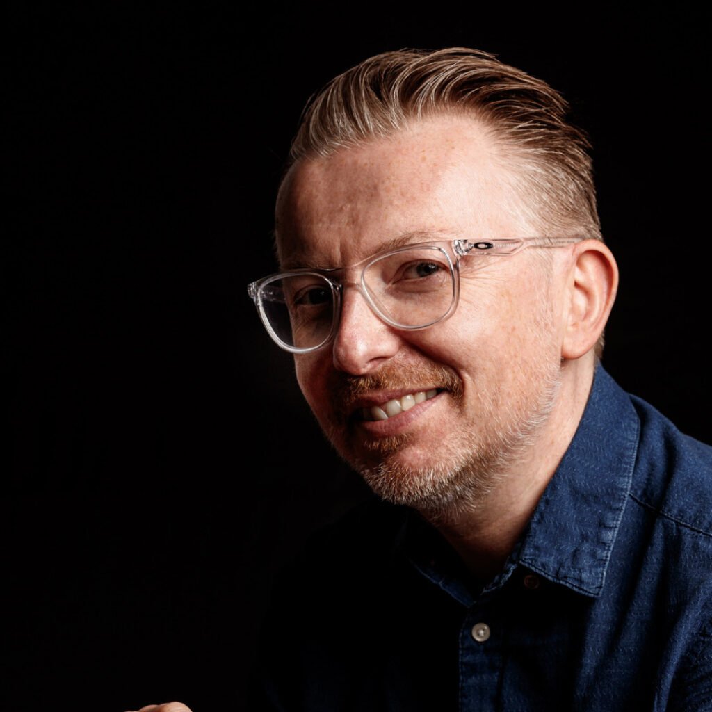 Headshot of Matt Peacock. He is wearing a blue shirt and glasses and is smiling. 