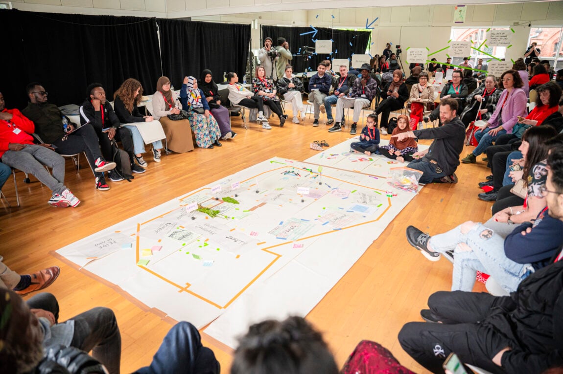 A room of people are gathered on chairs around 2 large pieces of paper on the floor that have notes and Post-its attached.