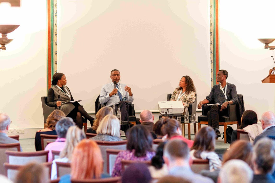 A panel of four people sit on a stage in conversations using microphones in front of a blurred audience. There is one black woman, two black men and one woman of mixed ethnicity.