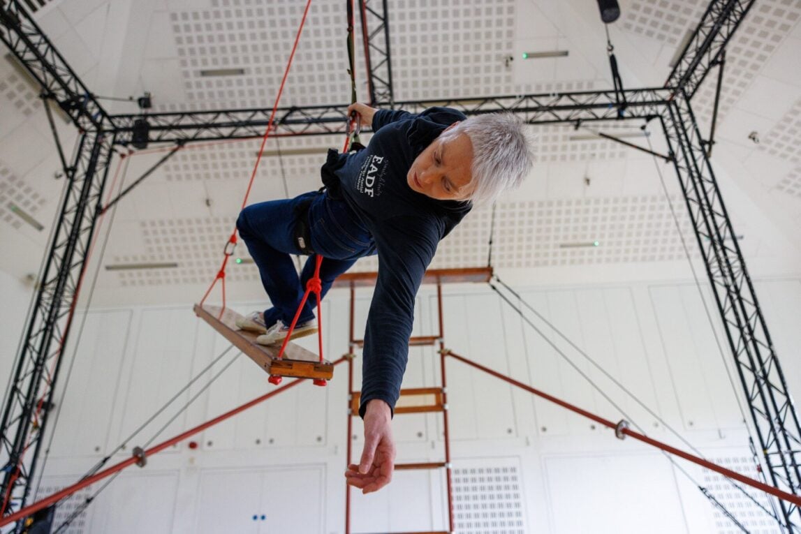 Lindsey Butler, a performer with short gray hair, is suspended mid-air on a wooden trapeze-like swing inside a white, high-ceilinged room with black scaffolding. Wearing a dark “EADF” sweatshirt and jeans, she leans dramatically to one side with one arm outstretched, appearing focused and graceful. The scene is part of an indoor aerial or physical theatre performance or rehearsal.